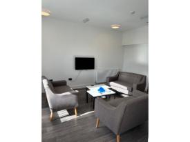 A brightly-lit lounge with sofas and armchairs arranged around coffee tables, facing a wall-mounted television. The room has white walls and large windows.