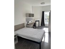 A bedroom with a double bed, study desk and chair, wardrobe, and a window with curtains. The room has light walls and dark flooring.