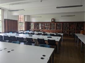 A large study room with tables and chairs in rows. Tall windows on one side and wooden bookcases filled with books along the back wall.