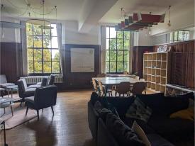 Lounge and study space with sofas, armchairs, a large table and chairs, tall windows, and shelving.