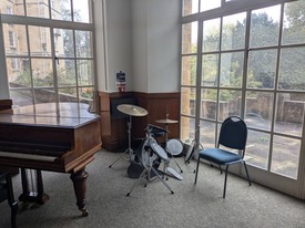A room with a grand piano and a drumkit. Large windows lookout onto a terrace and greenery.