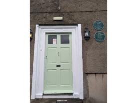 A pale green door set in a white frame on a textured wall. Two lanterns on either side, with two commemorative plaques to the right.