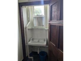 A white sink beneath a window, viewed through an open wooden door. The space has tiled walls, visible pipework, and a mirror above the sink.