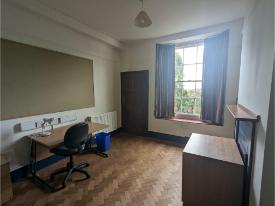A cosy room with a desk, office chair, and large window with curtains, letting in natural light. Wooden flooring and storage furniture line the walls.