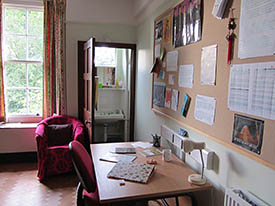 A room with a desk, office chair and armchair. There is a doorway into a small room containing a basin.