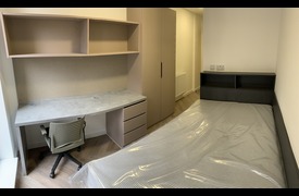 A small single bedroom with single bed, desk and chair, shelves and wardrobe.