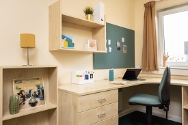 Corner desk and drawers with a chair. The room has a notice board above the desk and shelves with photoframe and potted plant.