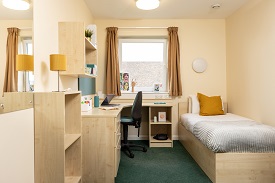 Single size bed to the right, a desk and chair to the left with a window and curtains to the back of the room.