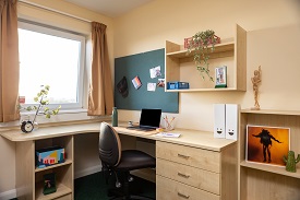 A wooden corner desk with drawers with a chair placed by the desk. The desk has a laptop, files, stationary and clock on it. Aobve desk is a pinboard and shelivng unit.