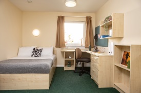 A double sized bed to the left of the image, corner desk with a chair  and drawers to the right. Aobve the desk is shelving and a pinboard. At the back of the room is a window with curtains.