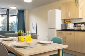 Wooden dining table at the forefront of the image dressed with juice, plates and cuterly. In the background is the wooden kitchen units and a fridge freezer.