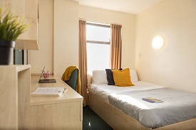A double sized bed to the right and a wooden desk with drawers and a chair to the left. At the back of the room is a window with curtains.