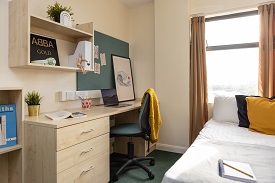 A double size bed to the right and a soft light on the wall. To the left is a desk with a chair.