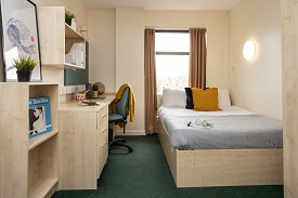 Bed and shelving units in the left foreground. Window and desk area in the background.