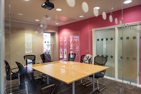 A room with glass windows with a large wooden table in the centre of the room and black chairs around the table.