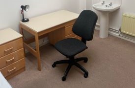 A room with a small chest of drawers, a desk and a black desk chair and a bathroom sink.
