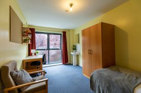 A room with a single bed against one wall, with a wardrobe at the foot of the bed and a sink with a mirror over it beyond it. On the opposite side of the room there is an armchair and a desk with an office chair next to it and a set of shelves on the wall above it.