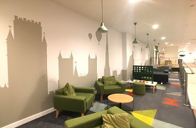 A room with several armschairs around a coffee table, a large Connect Four game, and some more armchairs and sofas beyond it.