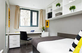A room with a bed against one wall and a desk with a chair against the adjacent wall. There is a set of shelves on the wall over the bed and a set of drawers under the desk.