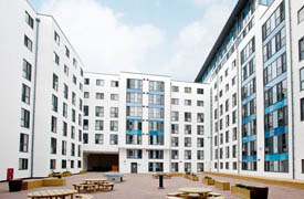 A large paved courtyard enclosed by a modern nine-storey building and containing several tables, benches, potted plants and a ping pong table.