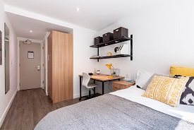 A room with a desk and a double bed against one wall. There is an office chair next to the desk and two shelves on the wall above it, and a wardrobe against another wall nearby.