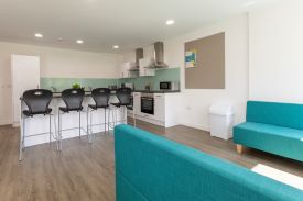 A kitchen with two ovens and hobs, two sinks, two microwaves and several cupboard and drawers. There are four tall chairs lined up against a kitchen counter and two sofas nearby.