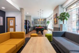 A large room with two sofas around a coffee table, two vending machines against a wall, and a pool table in the centre of the room. There is a high table with chairs around it at the far end of the room and the words '# the print hall' written in lights on the wall.