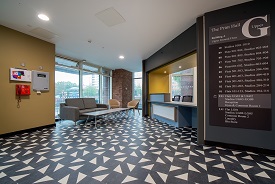 A large room with a reception counter and a sofa, coffee table and two armchairs nearby. The sign on the wall near the reception lists the buildings floors and shows that this is on the upper ground floor.