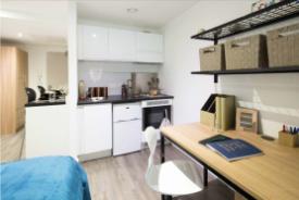 Studio flat with a kitchenette, study desk and chair, wall shelving with storage baskets, and part of a bed with a blue cover visible in the foreground.