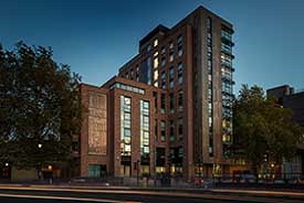 The exterior of a modern twelve-storey red brick building next to a road with traffic lights. The words 'the Print Hall MMXVII' are written on the side of the building.
