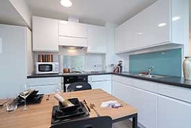 A kitchen with an over and a hob, a sink, a microwave and several cupboards and drawers. There is a dining table nearby with two chairs around it.