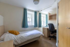 A room with a single bed in one corner and a desk against the opposite wall. There is an office chair next to the desk and a set of shelves above it.