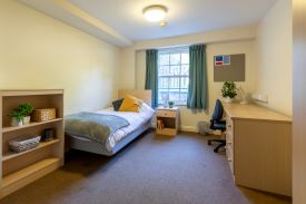 A room with a single bed in one corner and a desk against the opposite wall. There is an office chair next to the desk and a set of shelves at the foot of the bed.