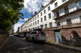 Exterior of a row of terraced multi-storey houses. There is a stairway leading up from the street to a raised pavement with iron railings.