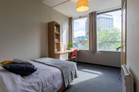 A room with a double bed and a desk in the corner at the foot of the bed. The desk has a chair next to it, a drawer below it, and a set of shelves against the wall above it.