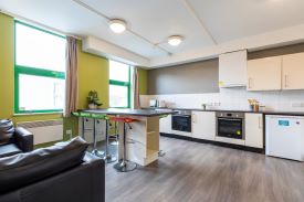 A kitchen with two ovens, a fridge and several cupboards and drawers against the walls. There is a separate kitchen counter with several stools next to it and two sofas nearby.