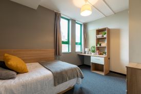 A room with a single bed in one corner and a desk agains the opposite wall. The desk has a drawer beneath it and shelves against the wall over it.