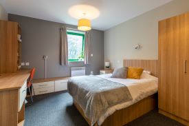 A room with a double bed and a wardrobe against one wall and a desk opposite. There is a chair next to the desk and several drawers and shelves against the walls around it.