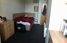 A room with a double bed in one corner and chest of drawers at the foot of the bed.