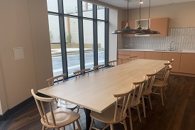 A large kitchen area with a long rectangular dining table with 13 chairs. On the far wall there is a sink, countertop and cupboard storage units. There is a large floor to ceiling window looking out on to a courtyard.