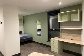 A bedroom with a double bed by the wall at the far end. At the opposite end there is a study area with a desk and shelving next to a window.