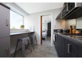 A kitchen with integrated appliances, breakfast bar seating by a large window, and a bedroom visible through a sliding door.