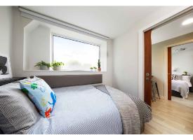 A bedroom with a double bed, large window, light wood flooring and a sliding door opening to an adjoining bedroom.