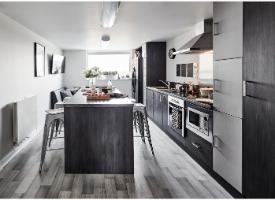 A modern shared kitchen with integrated appliances, a central breakfast bar, seating and a lounge area by the window.