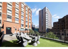 An outdoor rooftop terrace with seating, artificial grass and glass balustrades, overlooking nearby student accommodation buildings.