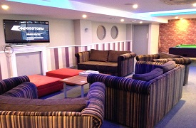 Social space, showing two sofas and three armchairs  gathered around a coffe table and a wall mounted screen. In the background there is a pool table