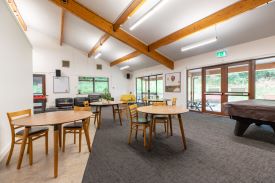 A large, well-lit common room with a vaulted ceiling. There are some tables and chairs, and sofas and armchairs. There is a pool table and a foosball table. There are some double doors that open out on to a patio area with some picnic benches.