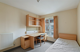 A naturally lit bedroom with a single bed. There is a desk, chair and shelves against the wall opposite the bed.