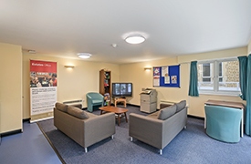 A common room with two sofas facing a coffee table and TV. There are two armchairs, a bookcase, and a printer/copier positioned by the walls.