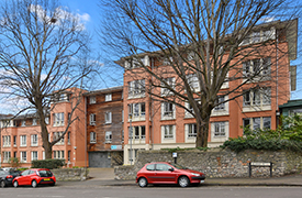 Street view of Woodland Court from Belgrave Road. The building is 5 stories high, and is positioned behind a brick wall and tall trees.